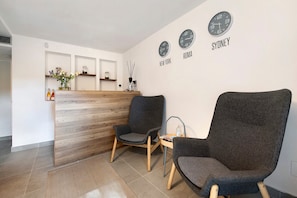 Zona de recepción con un mostrador de madera, dos sillones grises y relojes de pared con nombres de ciudades.
