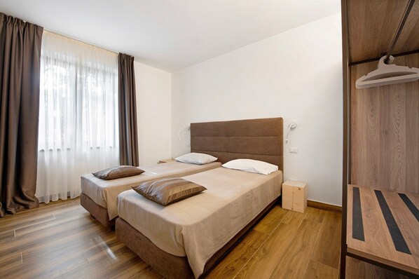 Una habitación con dos camas, suelo de madera, pared blanca y ventana con cortinas.