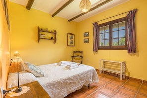 A bedroom with a bed, a bedside table with a lamp, a bench, a window with curtains, and a shelf with pictures. - Country House 'Lavanda' with Mountain View, Shared Pool and Wi-Fi (Órgiva)