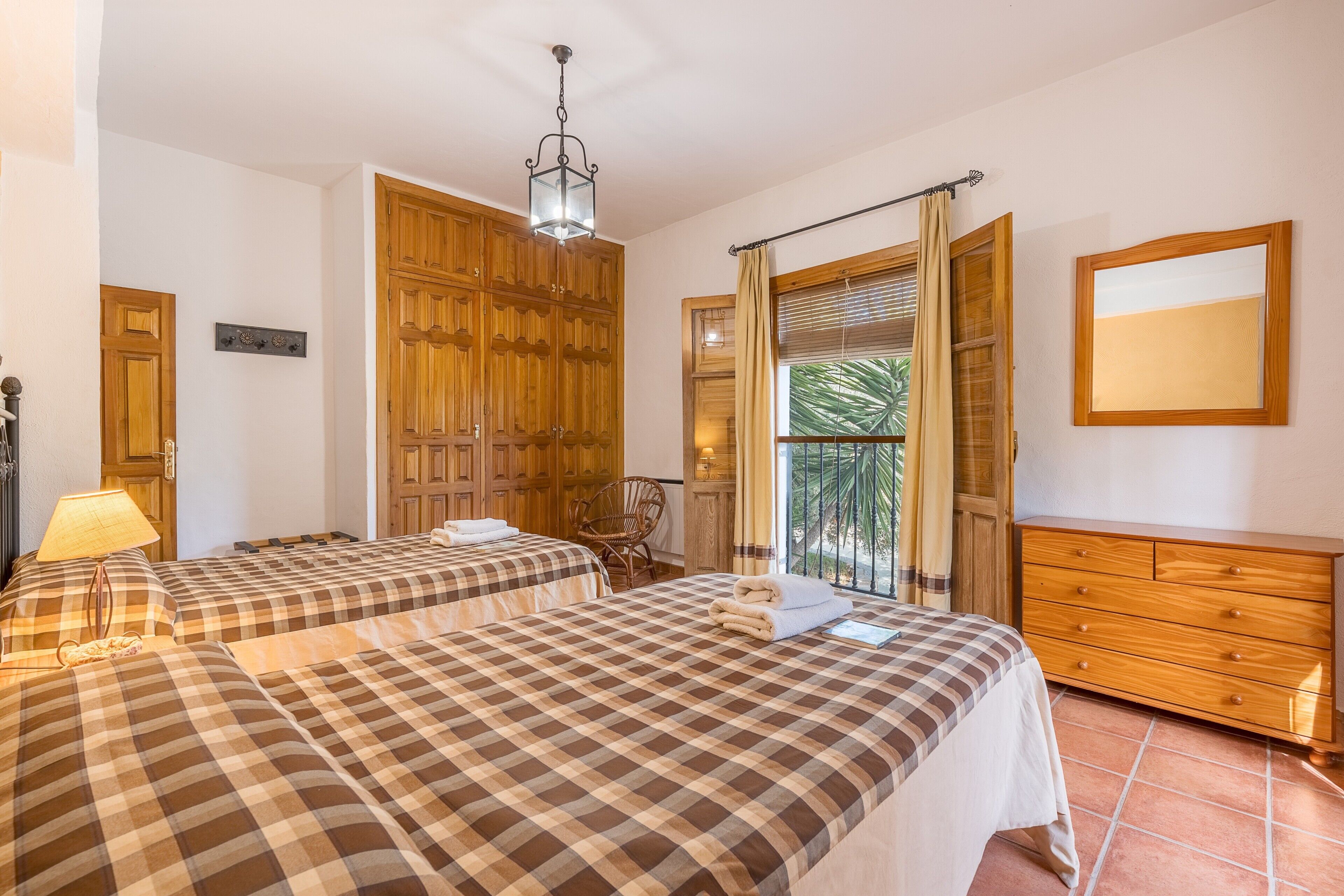 A hotel room with two beds, a wooden wardrobe, a dresser, a lamp, and a view of palm trees.