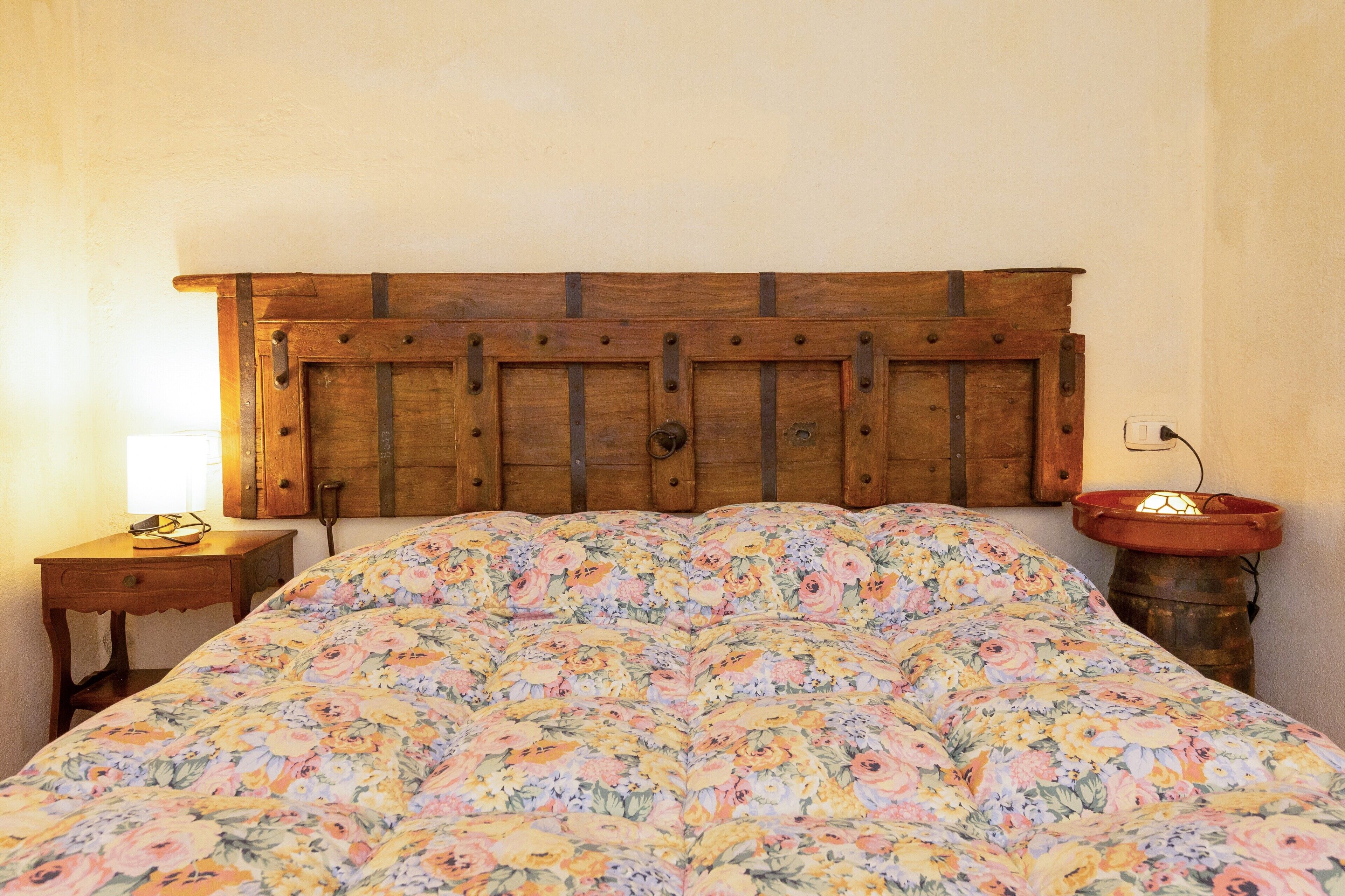 Un lit avec une couette à motifs floraux, une tête de lit en bois, une table de chevet avec une lampe et une table de nuit de style rustique.