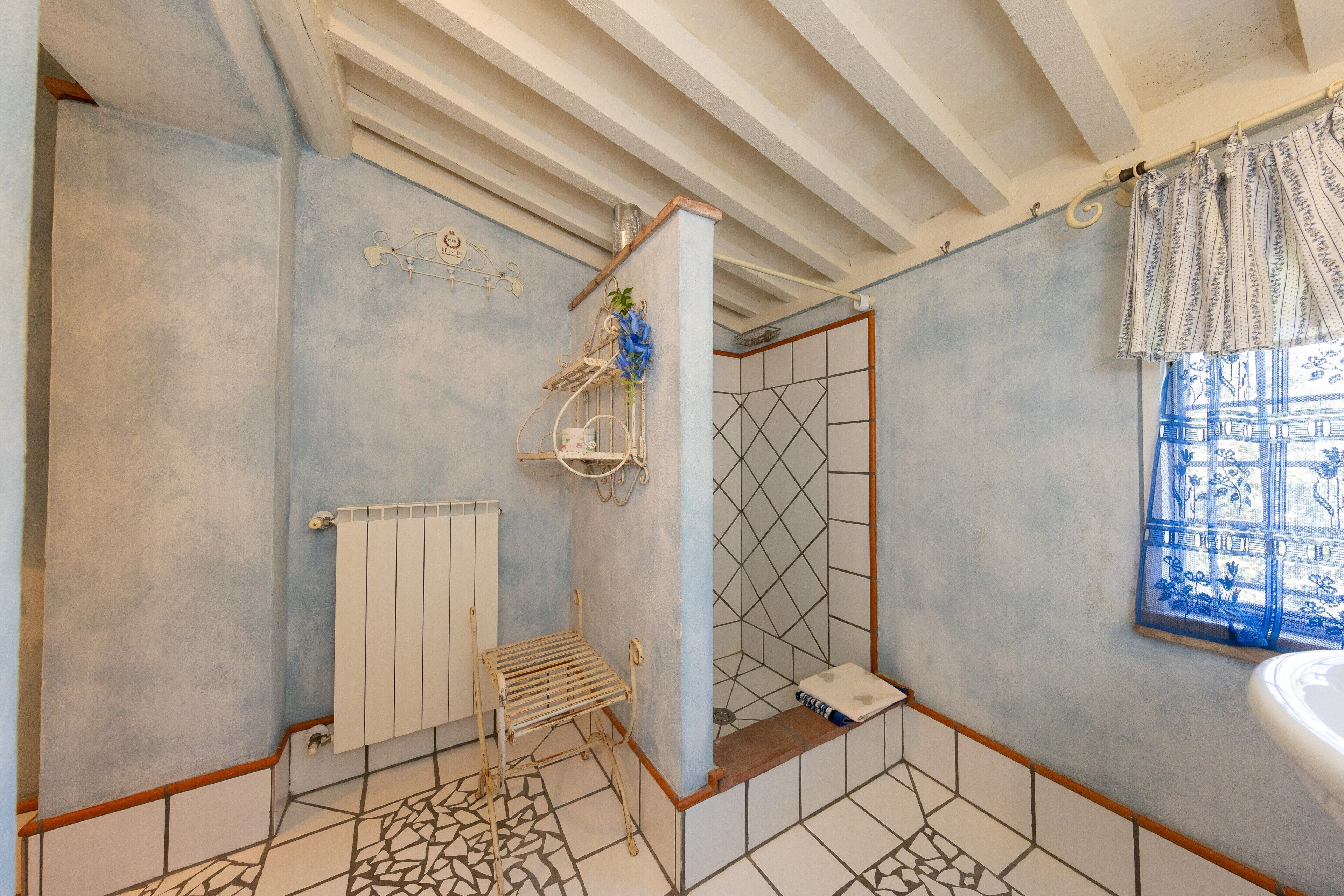 A bathroom with a tiled floor, a radiator, a shower with a geometric tile pattern, a window with a blue curtain, and a sink.
