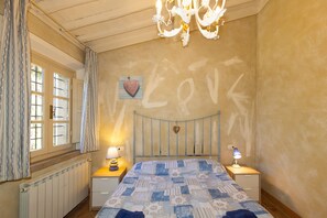 Une chambre à coucher avec un lit, deux tables de chevet, un lustre, une fenêtre avec des rideaux et un mur orné d’un décor en forme de cœur.