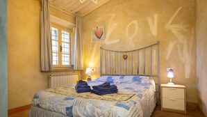 A bedroom with a bed, a nightstand, a lamp, a window with curtains, and a wall with a heart-shaped decoration. - San Giovanni R. Apartment 1 Dantea with Private Garden, Wi-Fi, and Air Conditioning (Peccioli)