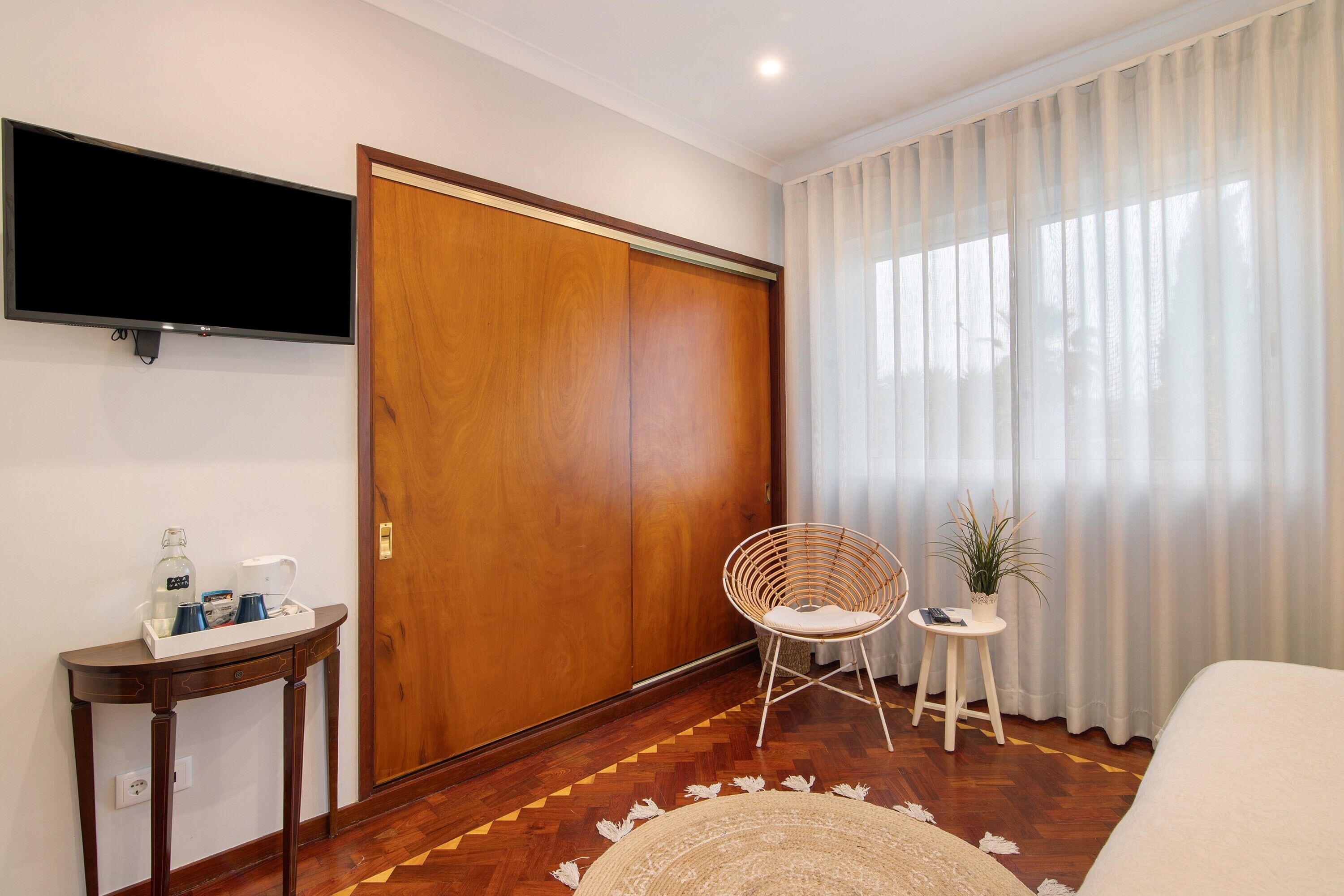 A hotel room with a wooden wardrobe, a television mounted on the wall, a small table with a plant, and a chair.