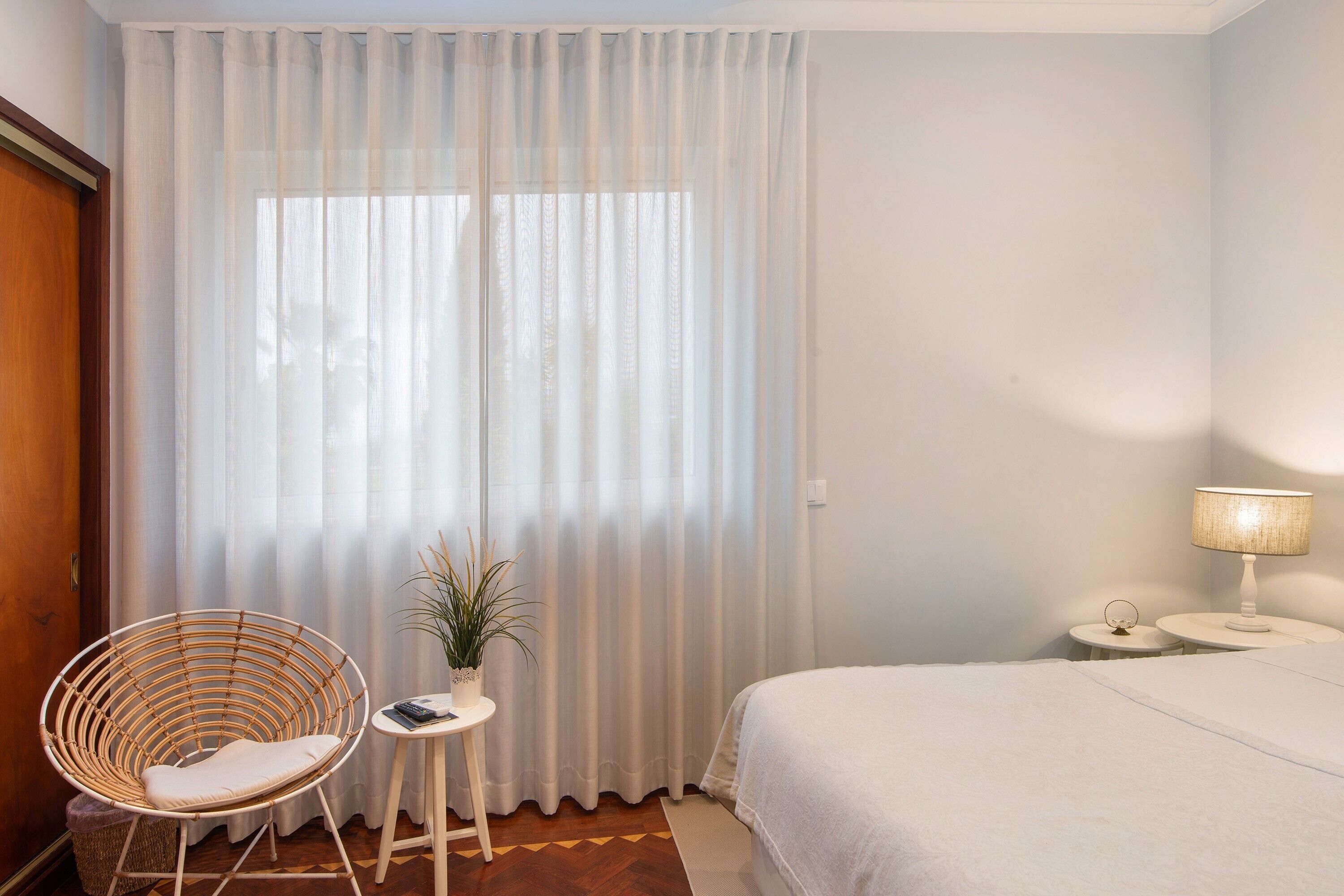 A room with a bed, a chair, a small table with a plant, and a lamp.