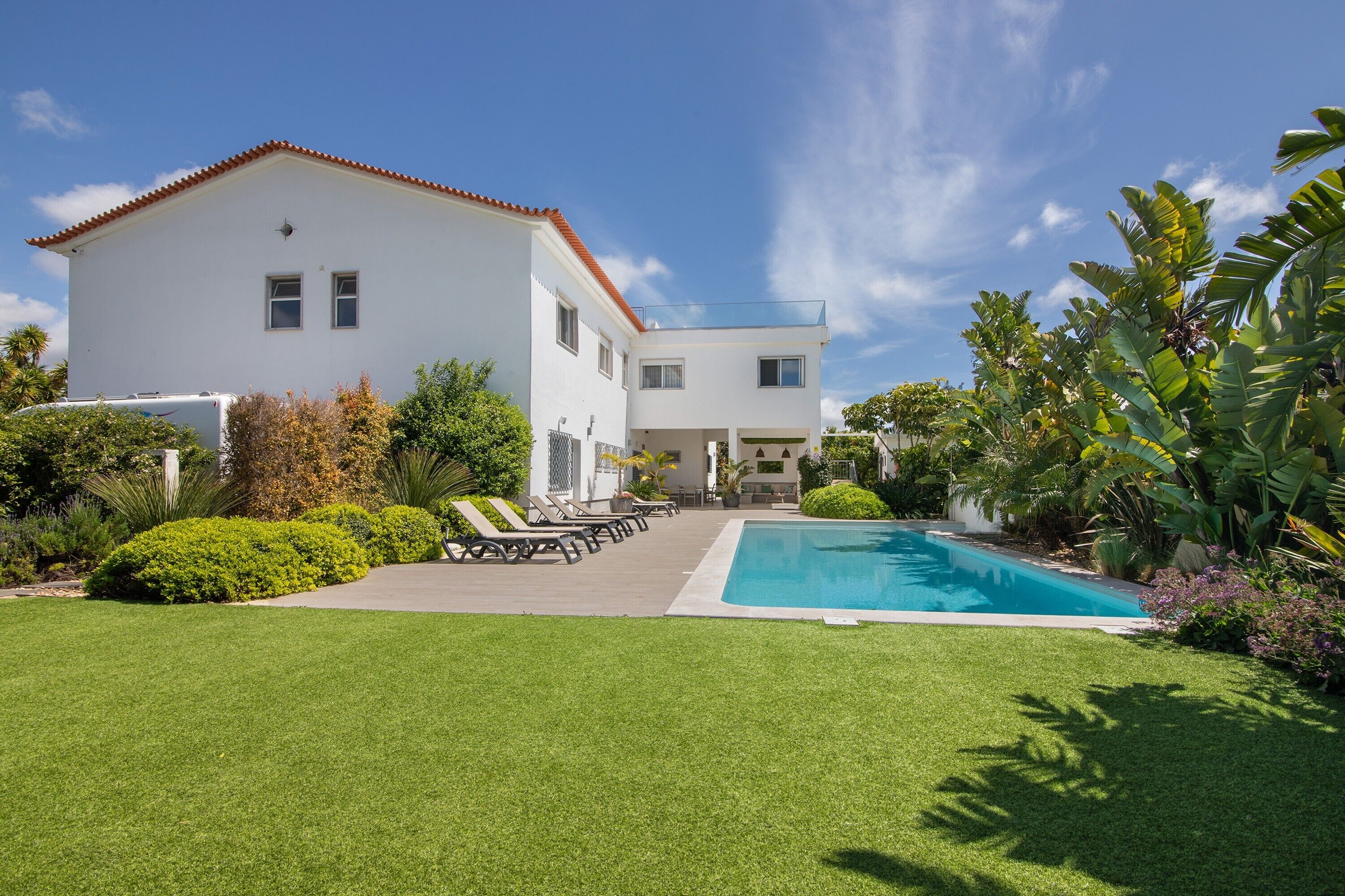 A two-story white building with a swimming pool, surrounded by well-maintained lawns and tropical plants.