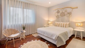A bedroom with a bed, a chair, a bedside table, a lamp, a wall decoration, and a window with curtains. - Milreu Guesthouse - Room "Raízes" (Estói)
