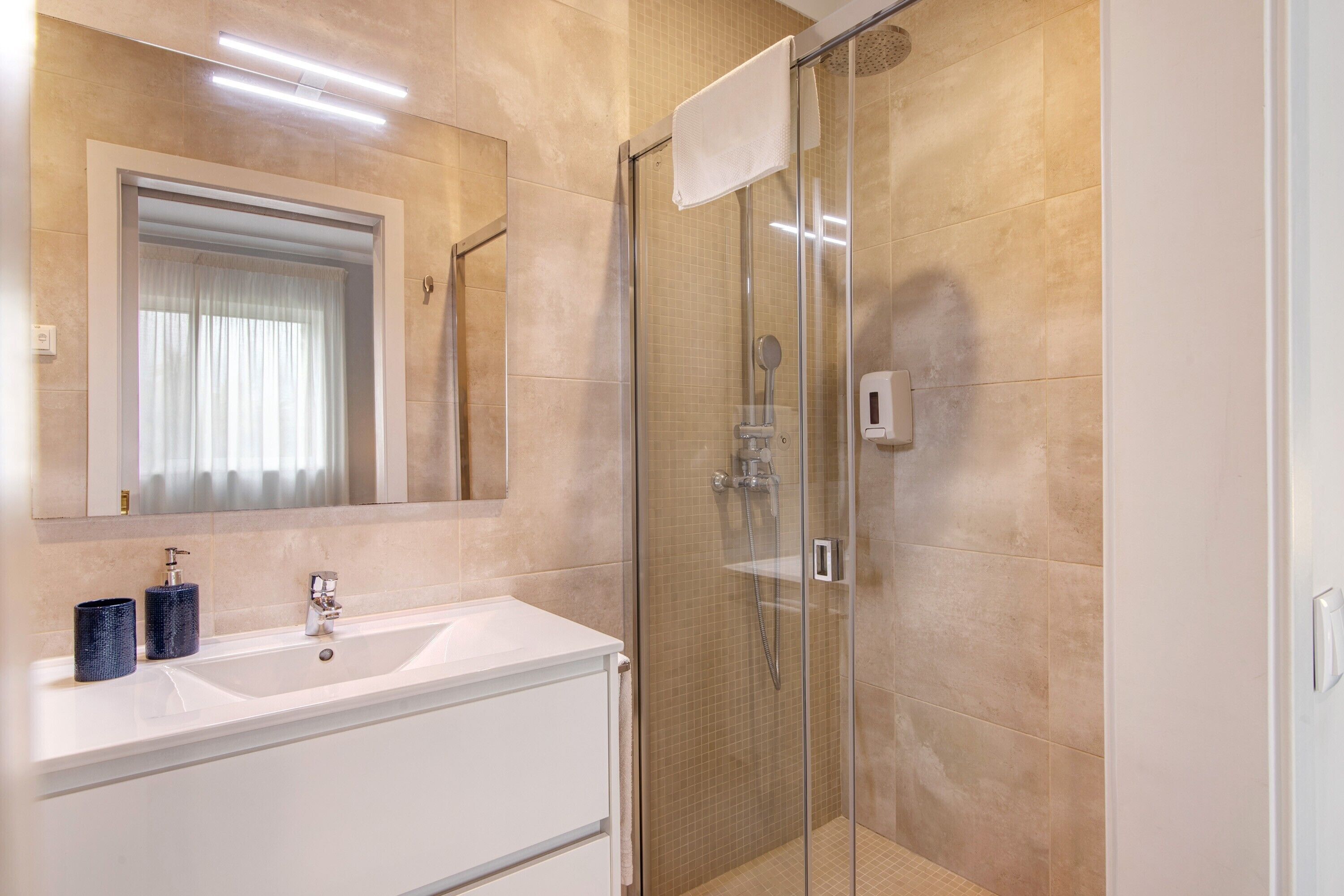 A modern bathroom with a glass-enclosed shower, a white sink with two bottles, and a large mirror above the sink.