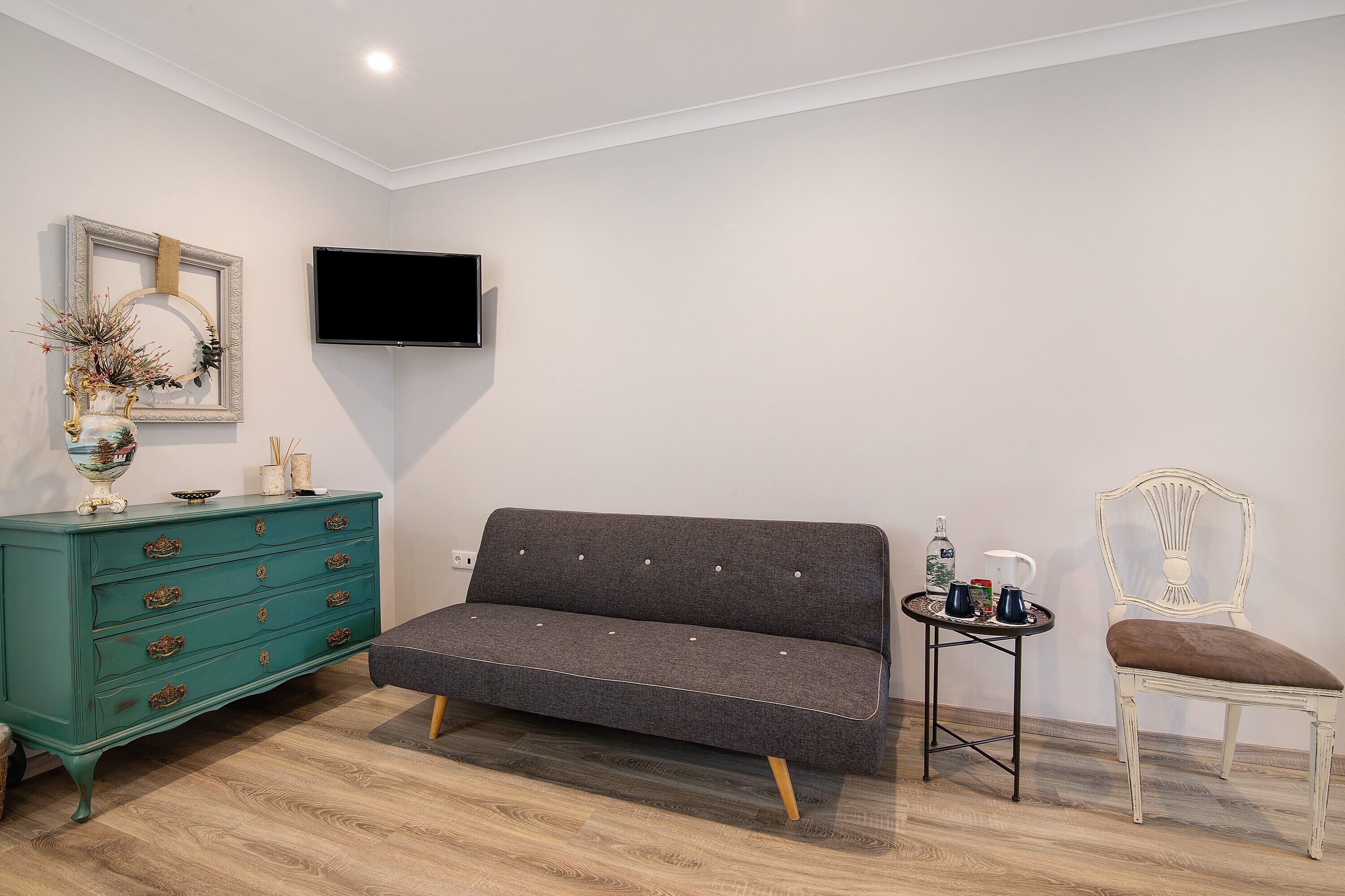 Une pièce avec un canapé gris, une commode vert turquoise, une chaise blanche, une petite table avec des boissons et un téléviseur fixé au mur.