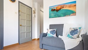 Una habitación con un sofá gris, una colcha blanca y un cuadro colgado en la pared que representa un paisaje costero.
