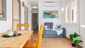 Una habitación con una mesa de comedor de madera, sillas, un sofá gris con almohadones decorativos, un cuadro colgado en la pared y una planta en maceta.