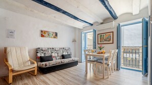Una sala de estar con sofá, una silla, una mesa de comedor con sillas, un balcón con barandilla y un cuadro en la pared.