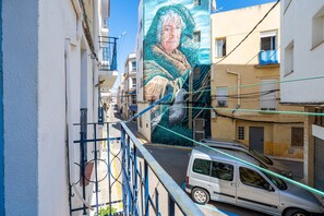 Una calle estrecha con un mural en un edificio, un coche blanco estacionado al lado y una barandilla azul.