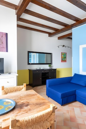 A room with a wooden ceiling, a TV mounted on the wall, a blue sofa, a dining table with a plate, and a kitchen area with cabinets.