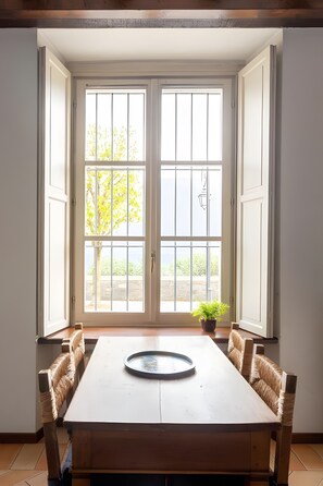 Un'area pranzo con un tavolo di legno e sedie in vimini, una pianta in vaso e una vista attraverso un'ampia finestra su alberi e un edificio.