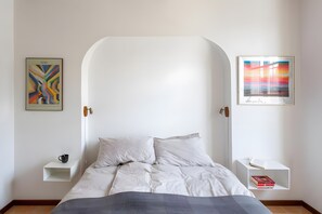 Ein Schlafzimmer mit weißem Kopfende, einem Bett mit weißer Bettwäsche, einem Nachttisch mit Tasse und Büchern sowie zwei gerahmten Kunstwerken an den Wänden.