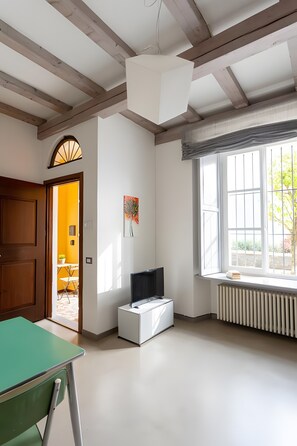A room with a wooden ceiling, a radiator, a window with blinds, a green table, and a door leading to another room.