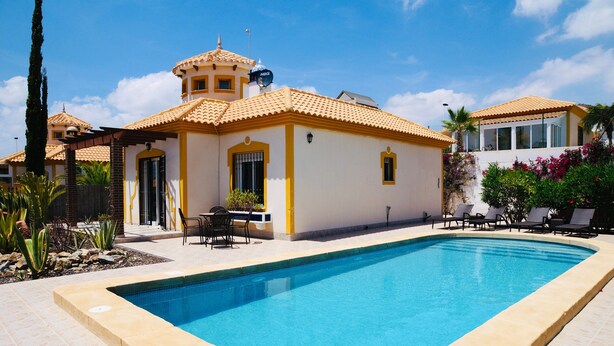 A swimming pool with a tiled border, a building with yellow and white walls, and a clear blue sky. - Villa 'Ensueno with Private Pool' with Private Pool, Wi-Fi and Air Conditioning (Mazarrón)