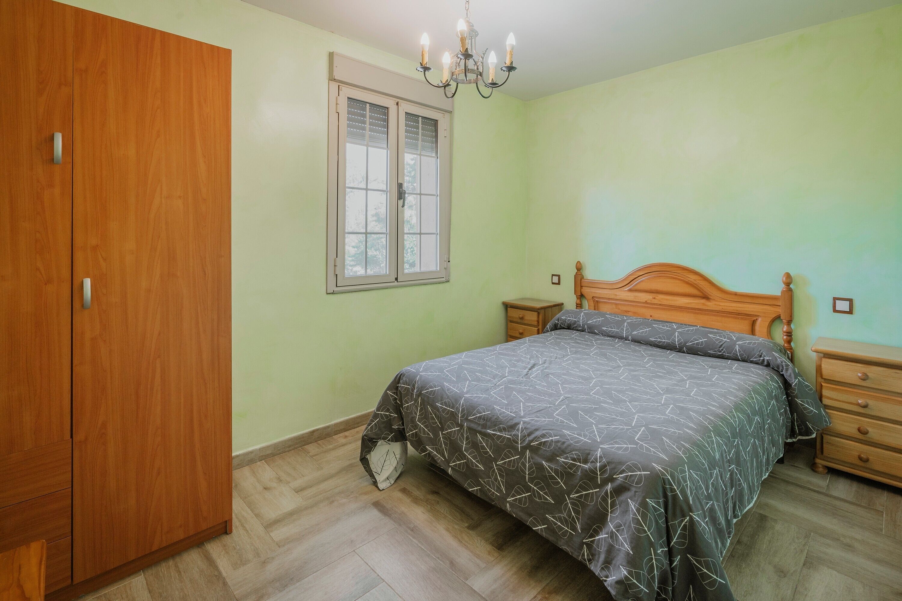 A bedroom with a wooden wardrobe, a bed with a patterned bedspread, a nightstand, and a window with blinds.