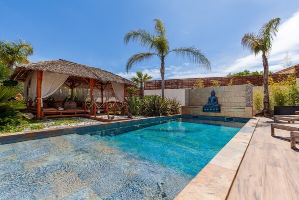 A swimming pool with a wooden deck, a thatched-roof structure, palm trees, and a Buddha statue.