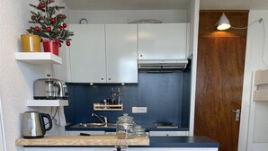 Una cocina compacta con gabinetes blancos, un pequeño árbol de Navidad y una mesa de comedor dispuesta con diversos objetos.