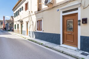 Vista de la calle con un edificio que tiene una puerta marrón, un letrero azul con el número 67 y una ventana pequeña con persianas.