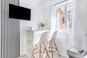 A modern room with a white desk, two white chairs, a potted plant, a TV, and a window with curtains.