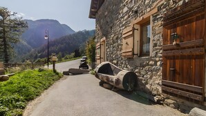 Un edificio de piedra con contraventanas de madera, un muro de piedra y un camino pavimentado que lleva a una montaña.