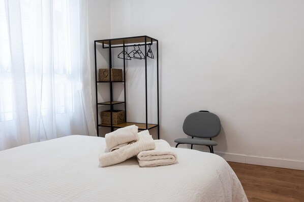 A neatly made bed with folded towels, a black metal shelf with baskets, and a gray chair in a room with white walls and wooden flooring.