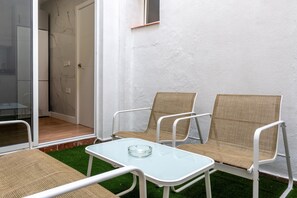A room with a glass table, two wicker chairs, a small glass ashtray, and a view of a door and a wall with a textured surface.