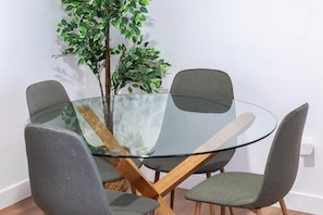 A round glass table with four grey chairs, a wooden crossbar, and a green plant in the background.
