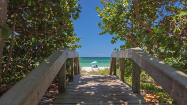 Ein Holzsteg führt zu einem Strand, wo eine Person unter einem Sonnenschirm entspannt.