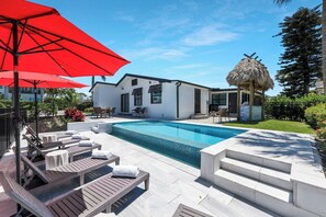 A modern house with a swimming pool, outdoor seating, and a thatched-roof structure. - Canal Dreamin | Heated Pool • Mini Golf • Waterfront Oasis Near Vanderbilt Beach (Naples)