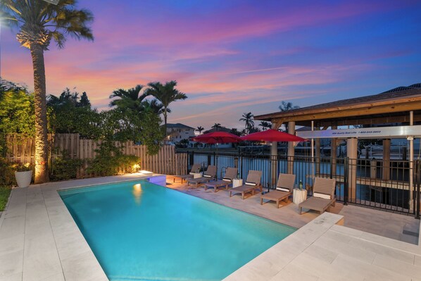 A swimming pool with lounge chairs and umbrellas, surrounded by palm trees and a wooden fence. - Canal Dreamin | Heated Pool • Mini Golf • Waterfront Oasis Near Vanderbilt Beach (Naples)
