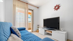 Una sala de estar con un sofá, un televisor de pantalla plana, un mueble blanco para entretenimiento, una decoración de timón rojo en la pared y una puerta corrediza de vidrio que da a un balcón exterior.