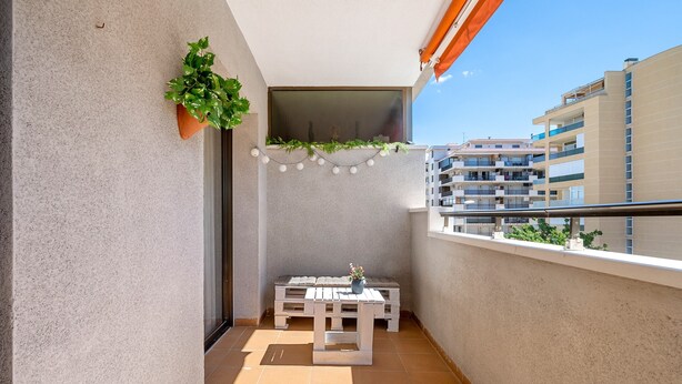 Un balcón con una mesa blanca, plantas en macetas, luces colgantes y vistas de edificios cercanos.