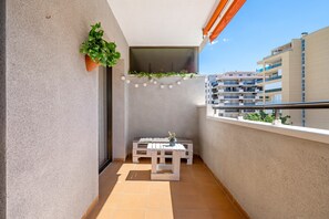 Un balcón con una mesa blanca, plantas en macetas, luces colgantes y vistas de edificios cercanos.