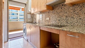 Cocina compacta con armarios de madera, encimera de granito, lavadora y ventana con persianas.