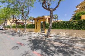 Una calle con un edificio amarillo identificado como 'Las Arenas', rodeado de árboles y un seto.