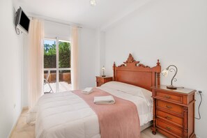 Une chambre à coucher avec un lit, une table de chevet en bois, un téléviseur fixé au mur et une fenêtre avec des rideaux.