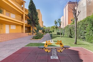 A playground with seesaw and swings, a yellow building with balconies, and a pathway lined with trees. - Apartment 'Las Arenas' with Shared Pool, Private Terrace and Wi-Fi (Islantilla)