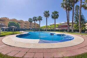 A swimming pool with a surrounding walkway, palm trees, and hotel buildings in the background. - Apartment 'Las Arenas' with Shared Pool, Private Terrace and Wi-Fi (Islantilla)
