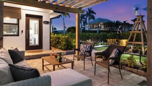 A covered outdoor patio with a wooden pergola, seating area with a sofa and chairs, a small table with drinks, and a view of a golf course and palm trees at dusk. - 🏖️ Coastal Luxury Retreat | Private Pool & Spa • Walk to Vanderbilt Beach • Naples Park Gem (Naples)