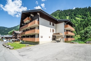 A multi-story hotel with balconies, a parking area, and a mountainous backdrop.