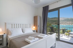 Une chambre moderne comprenant un lit, des tables de chevet, une commode, un balcon avec une table et des chaises, et une vue sur un plan d’eau et des collines.