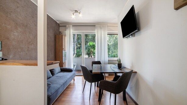 A modern living room with a sofa, dining table, chairs, and a TV mounted on the wall. - Apartment 'Modern Park' with Shared Garden, Balcony and Wi-Fi (Meran)