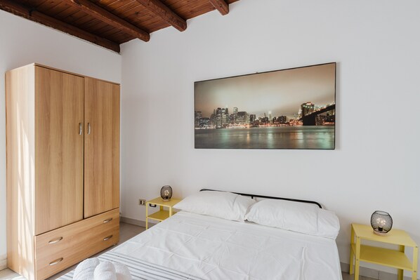 A bedroom with a bed, a wooden wardrobe, a nightstand with a lamp, and a cityscape painting on the wall.