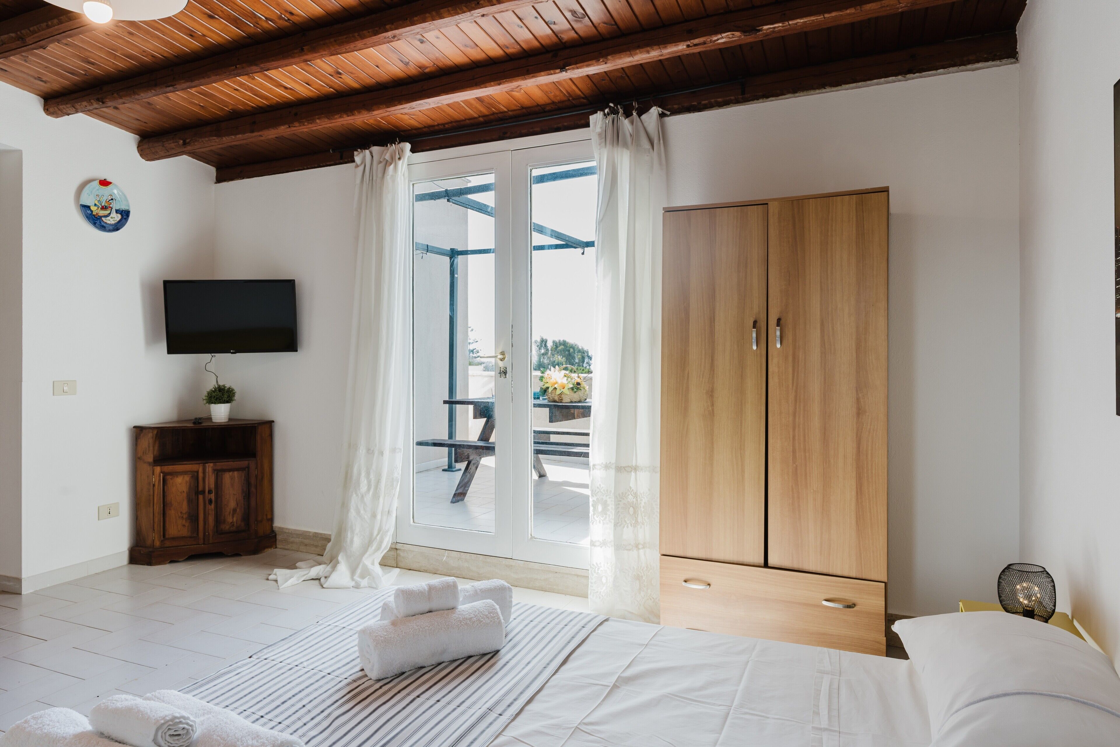 Een slaapkamer met een houten plafond, een televisie aan de muur, een kledingkast en een raam met gordijnen.