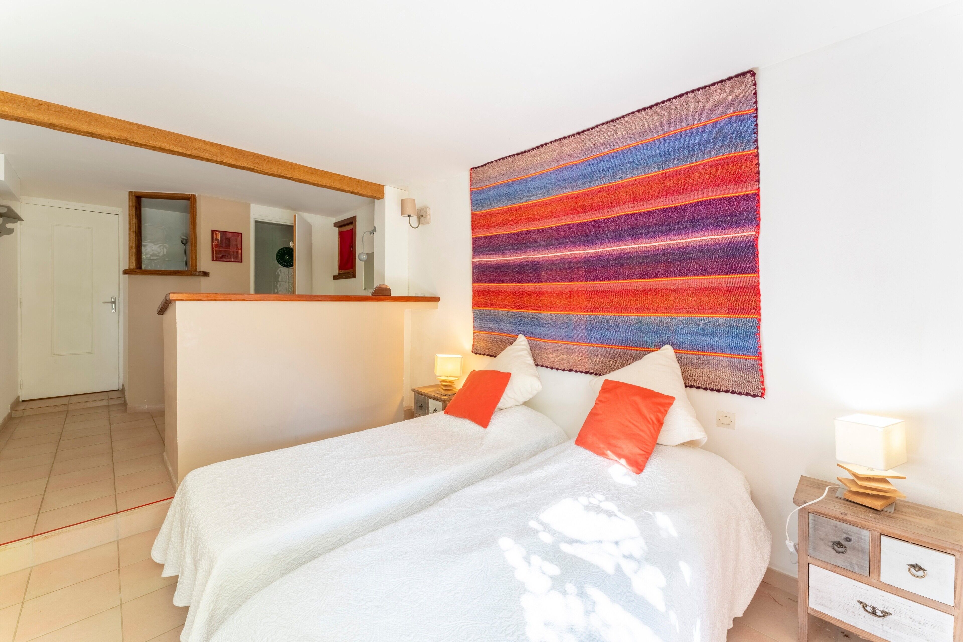 Ein Zimmer mit einem Bett, einem Nachttisch, einem Spiegel und einem farbenfrohen Wandteppich.